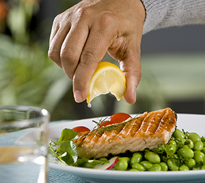 Closeup of man's hand squeezing lemon on broiled salmon.
