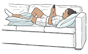 Woman lying on sofa with ice pack on bandaged knee and leg elevated on pillows.