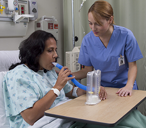 Proveedora de atención médica mostrándole a una mujer que está en cama en el hospital cómo usar un espirómetro de incentivo.