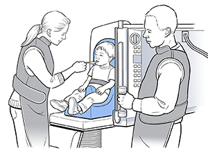 Small boy sitting in child seat on X-ray machine. Health care provider is feeding boy from spoon. Another health care provider is standing nearby.