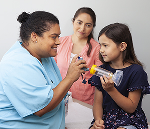Una proveedora de atención médica le muestra a una niña cómo usar un inhalador de dosis medida con un espaciador y una máscara. Mujer parada cerca.