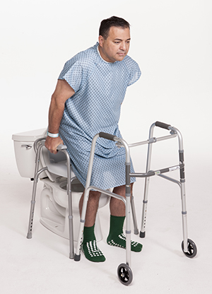 Hombre con una bata de hospital en el sanitario que se sostiene de las barandillas laterales del inodoro para mantenerse firme mientras se sienta en el inodoro.