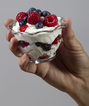 Mano sosteniendo un plato de vidrio con yogur y bayas frescas.